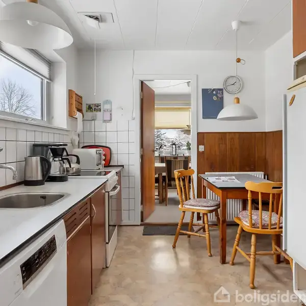 Køkkenet har hvide fliser, en hvid vask og træskabe. To stole står ved et bord med ur og lampe over. Vinduet giver udsigt til træer.