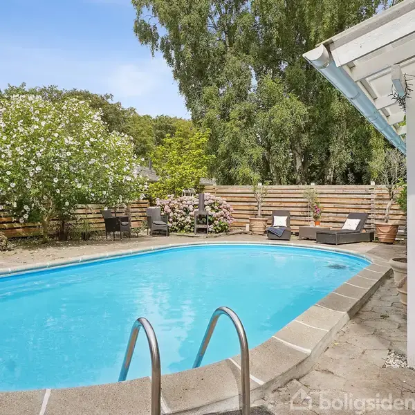 En swimmingpool med stige står stille i en flisebelagt have. Omkring poolen er der havemøbler, blomstrende buske og en pergola i skyggen af træer.