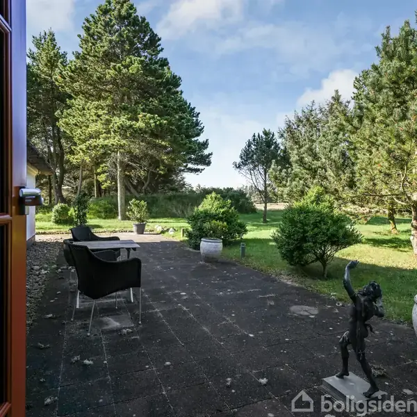 Terrasse med sorte stole på flisebelagt område, omkranset af grønne buske og store træer, set fra en glasdør, under blå himmel.