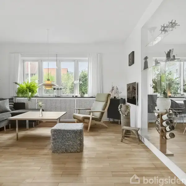 Stue med grå sofa og lænestol, arrangeret omkring et sofabord. Store vinduer giver naturligt lys og har planter i karmen. Til højre ses et spisebord med stole i et åbent rum.