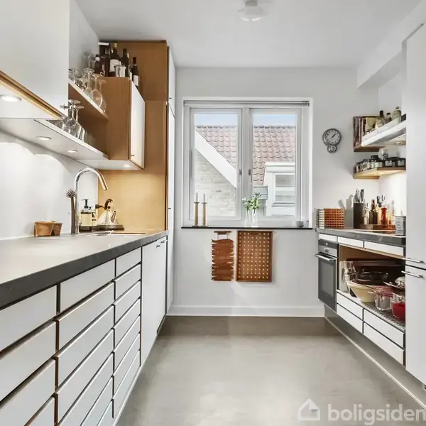 Et moderne køkken med hvide skabe og trædetaljer. Skabene er fyldt med krydderier og køkkenværktøj. Et vindue for enden lukker dagslys ind.