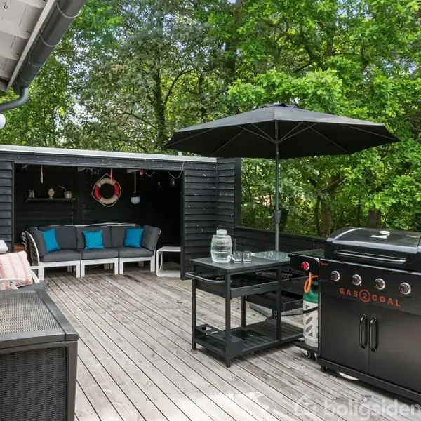 Terrasse med gasgrill placeret ved et bord med parasol. I baggrunden står en bænk med farvede puder under et halvtag. Omgivet af grønne træer.