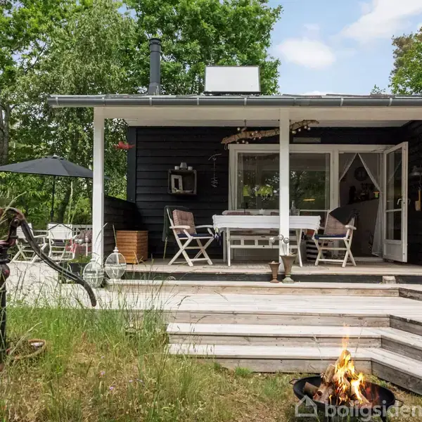 Sort træhus med åbne døre, hvide udendørsmøbler og terrasse i skovomgivelser. Foran brænder et lille bål i en skål i græsset.