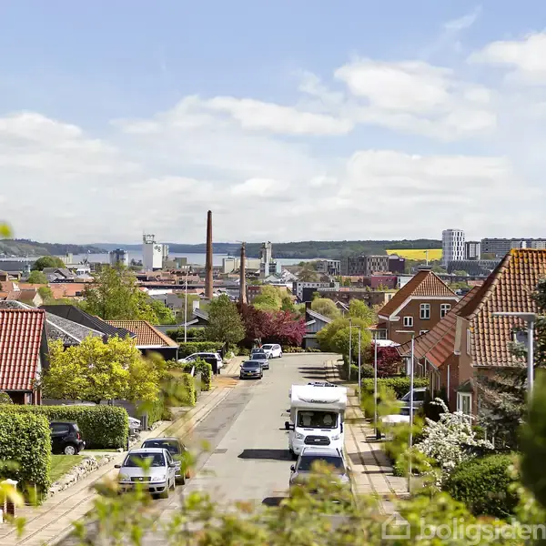Rækkehuse med røde tage står langs en stille vej, hvor biler er parkeret. I baggrunden ses en byskysline med industri og grønne områder under en blå himmel med skyer.