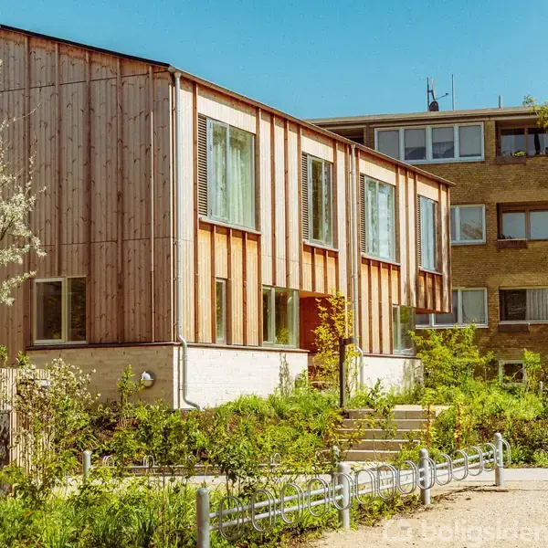 Et moderne træbeklædt rækkehus med store vinduer står i en grøn have med blomster og buske. I baggrunden er der en ældre lejlighedsbygning med flere etager.