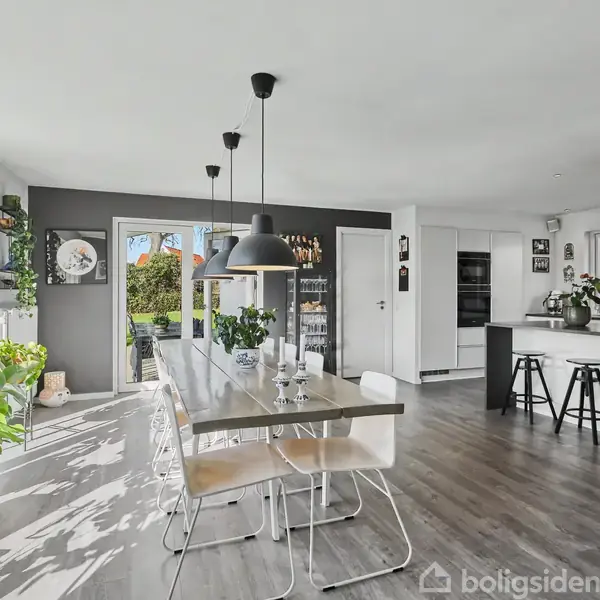 Moderne spisestue med langt bord og hvide stole, omkranset af vinduer og grønne planter. Sort lampe hænger over bordet. I baggrunden ses et køkken med barstole og hvide overflader.