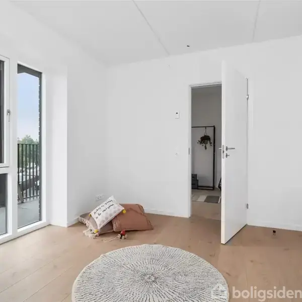 Et lyst værelse med en åben dør, trænger til indretning. En rund tæppe ligger på gulvet, puder er stablet langs væggen. Store vinduer giver udsigt til en balkon.