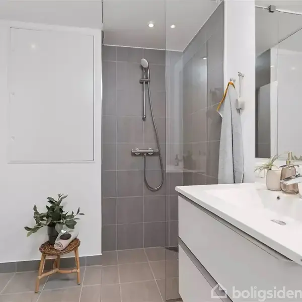 Badeværelse med grå fliser, installation af brusehoved og glasvæg. Hvid håndvask med spejl, og lille plante står på stol ved toilet. Enkel og moderne indretning.
