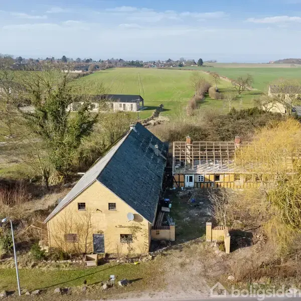 En gulstenslade med skråt tag står forladt og forfaldent. Huller i taget afslører forværet. Omgivet af åbne marker og småbygninger under en klar himmel.