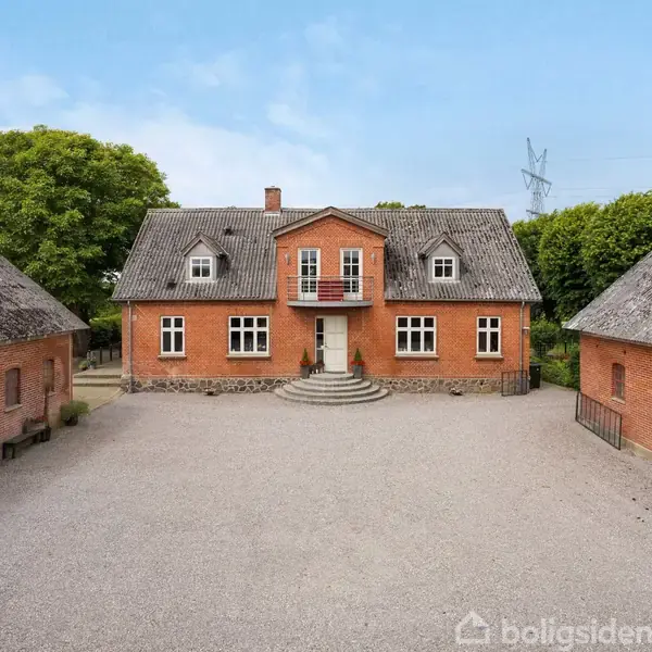En gård med en hovedbygning omgivet af grønne marker og træer. En vindmølle står tæt på bygningerne. Landskabet omkring er fladt og landligt med elektricitetstårne i baggrunden.