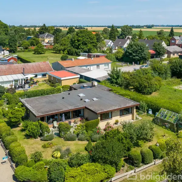 En moderne bungalow med fladt tag er omgivet af en velplejet have. Den ligger på en rolig, villavej i et grønt, landligt område med flere huse i baggrunden.