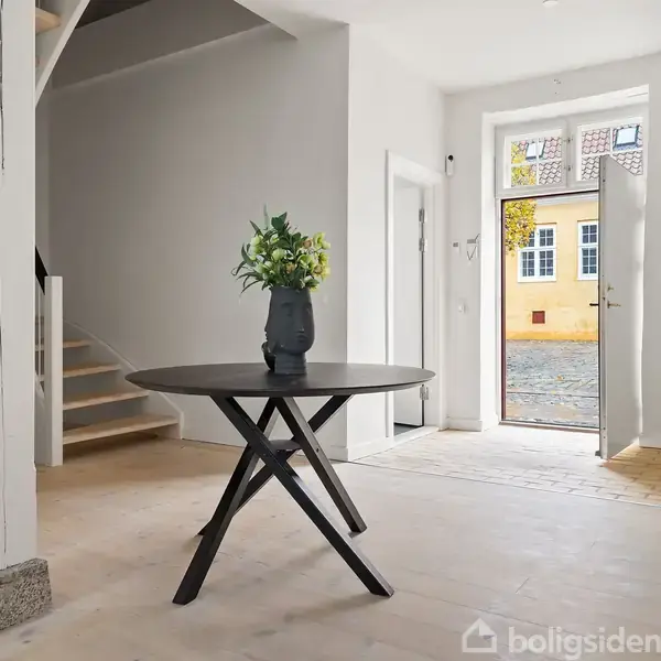 Et rundt bord med en vase med blomster står i en lys gang. En åben dør fører ud til en brostensbelagt gade og gule bygninger.