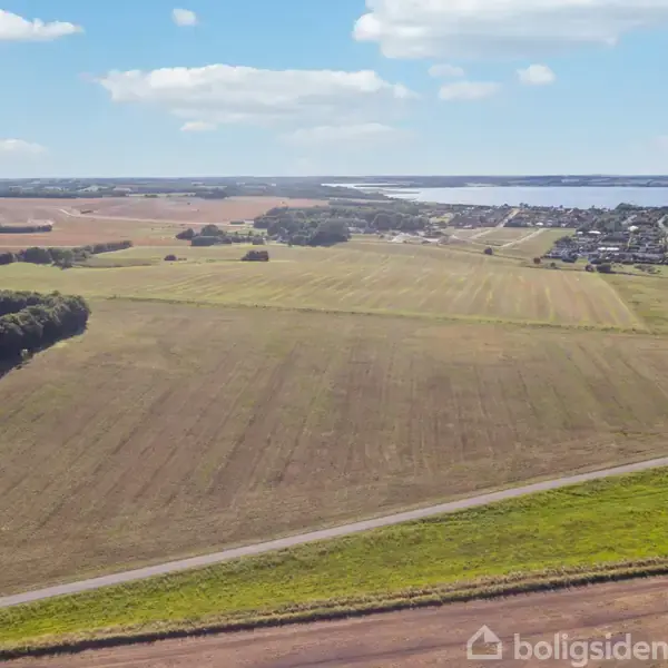 Store marker strækker sig over et landskab; en vej skærer gennem markerne. I baggrunden ses en landsby og en bred sø, omgivet af skyer på en klar dag.