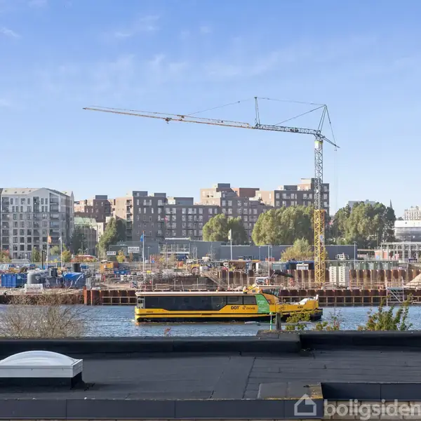 En pram sejler på en kanal foran en byggeplads med en kran og bybygninger i baggrunden. Himlen er blå, og der er træer langs vandet.