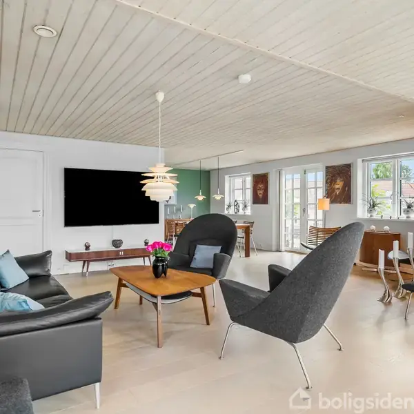Stue med sorte møbler. En sort sofa og to stole står omkring et træbord med blomster. Kunst pryder væggene. Store vinduer slipper lys ind. Fernisagtigt loft og gulv.