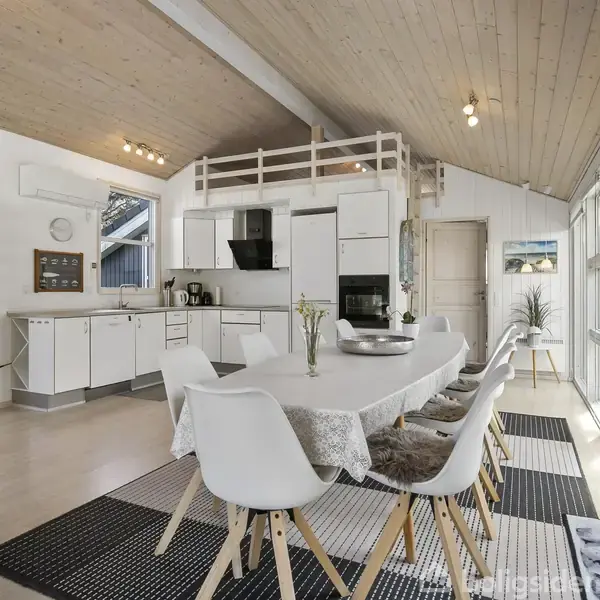 Spisebord med hvide stole står i et lyst køkken med hvide skabe. Ovenpå er der en hems. Store vinduer til højre giver udsigt til naturen udenfor.