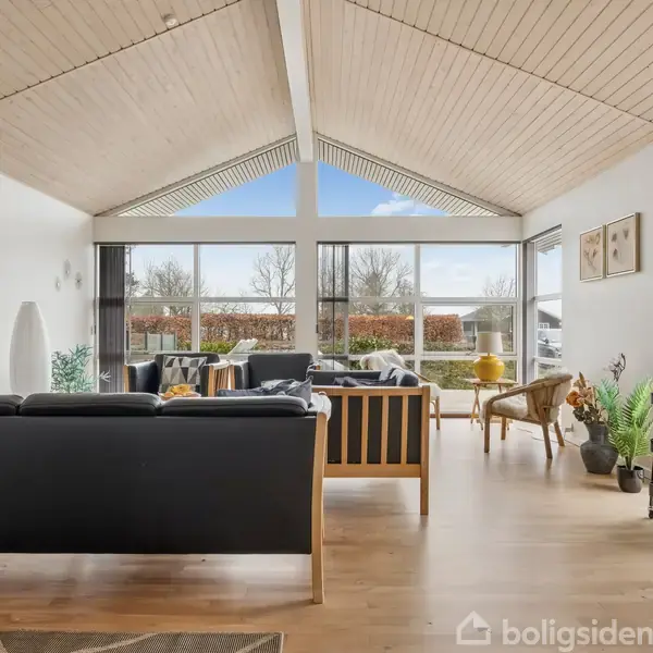 Stue med højt loft og store vinduer, indeholdende sorte sofaer og et sofabord. Plante og en lampe står ved siden af, med udsigt til en have.