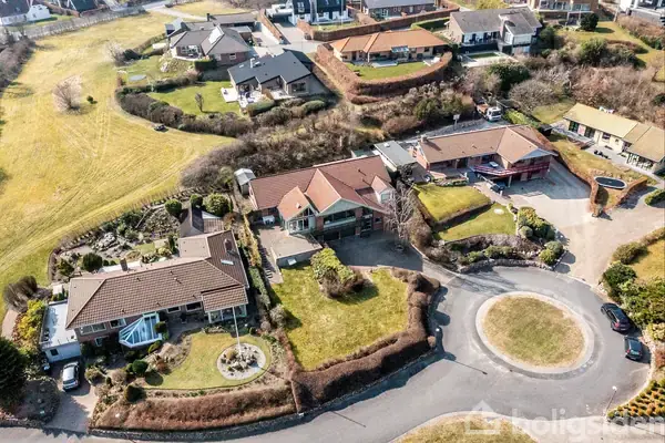 Luftfoto viser en gruppe huse med skrånende tage, omgivet af velplejede haver og grønne områder. De ligger i et boligområde med cirkulære veje.