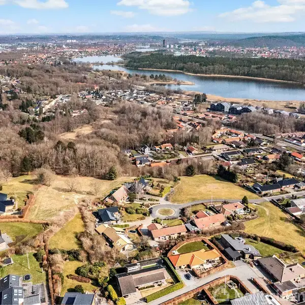 Dronebillede viser en forstad med spredte huse, marker og skove i forgrunden. En stor sø og en by i baggrunden under en klar, blå himmel.