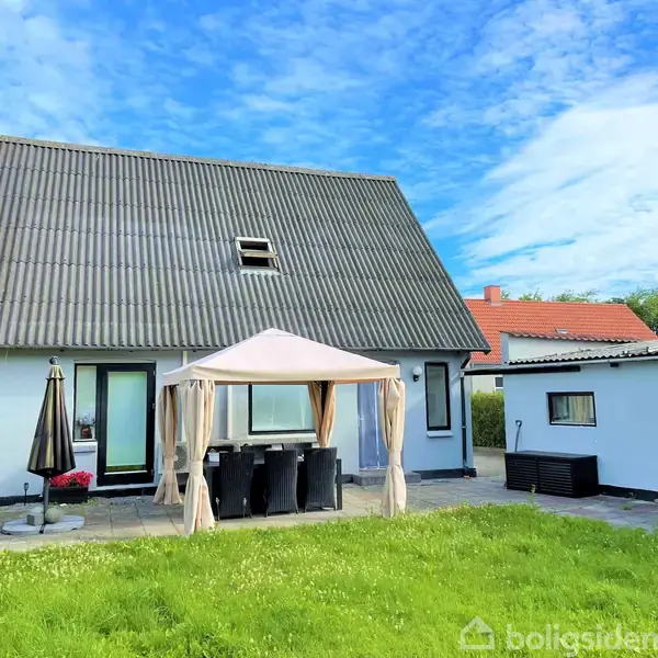Et hvidt hus med mørkt tag står i en velplejet have. En pavillon beskytter havemøbler på terrassen. Baghus ved siden af, under blå himmel med få skyer.