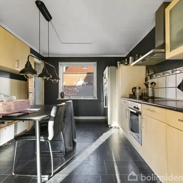Køkken med lyse træskabe og sorte fliser, bord med stole i midten, vindue bagerst viser hustage. Lyspendler hænger over bordet. Køkkenmaskiner på bordpladen.