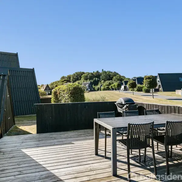 En træterrasse indeholder et sort spisebord med seks stole og en grill. Terrasse omgivet af et sommerhusområde med grønne buske og klare blå himmel.