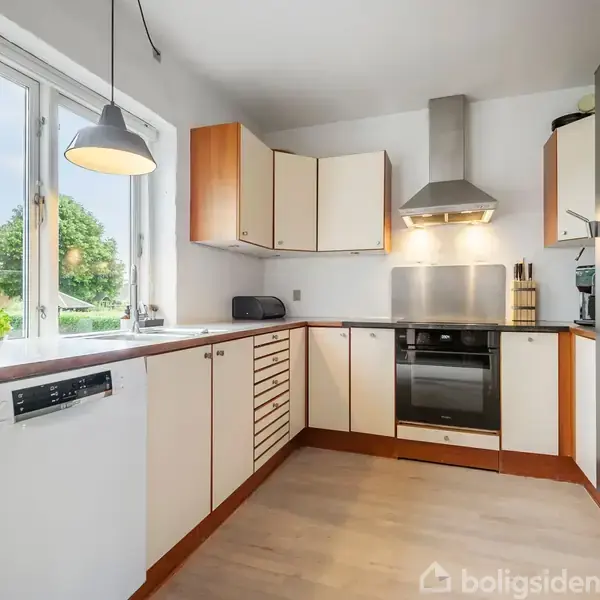 Stue med grå sofa og lænestol ved siden af et rundt sofabord med vase og lys. En brændeovn er i hjørnet, og en åbning leder til spisestuen med spisebord.