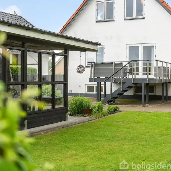 Et sort træhus med store vinduer og en overdækket terrasse, hvor der hænger en hængekøjestol. Terrassen er omkranset af have med græsplæne og planter.
