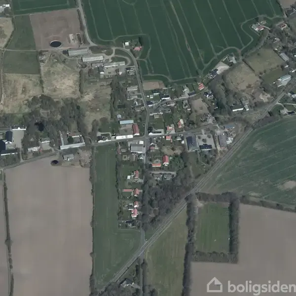 Luftfoto viser en landsby med huse langs en vej omgivet af marker og grønne marker. Skovområder ses i baggrunden, mens beboelsesstrukturer er centralt placeret.