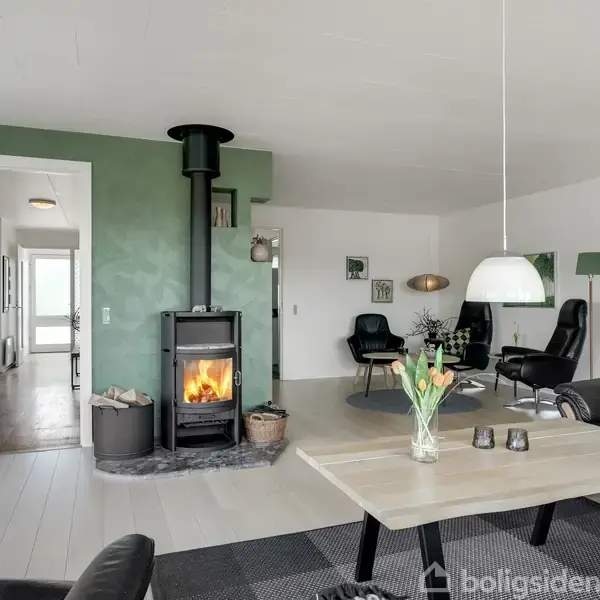 Brændeovn opvarmer stue med grøn væg; spisesofabord med tulipaner; læderlænestole ved siden af gulvlampe; indgang til gang med synligt lys.