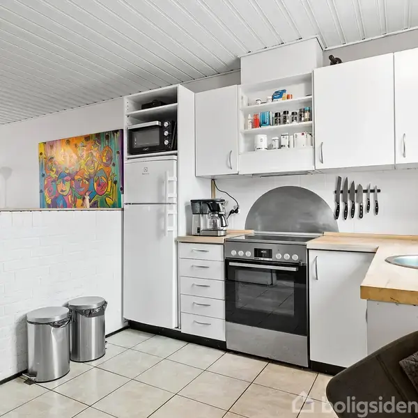 Et moderne køkken indeholder et komfur og ovn, med knive på væggen og opvask ved vasken. Køleskab, mikroovn, og skabe omgiver det. Et kunstværk hænger i stuen ved siden af.