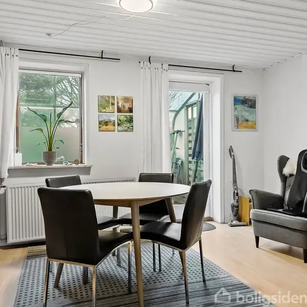 Et rundt spisebord står med fire stole omkring i en lys stue med trægulv. En reol fyldt med bøger er placeret ved siden af vinduer med hvide gardiner. En grå lænestol står tæt på en væg.