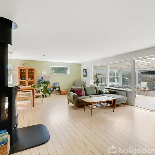 En moderne stue med en fritstående sort brændeovn til venstre. Et sofabord og en grøn sofa står mod højre. Store vinduer åbner mod en terrasse. Reolkasse og planter til venstre.