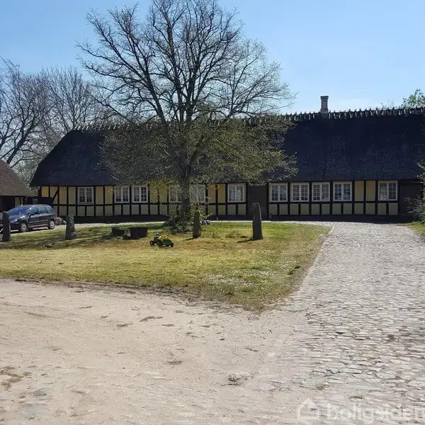 Bindingsværkshus med stråtag står i solskin, omgivet af græsplæne og brostensbelagt gårdsplads. En bil er parkeret til venstre, og et stort træ står foran huset.