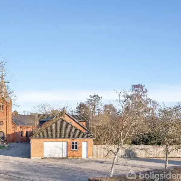 En lille, rød murstensbygning med skråt tag står foran en større kirke med et højt klokketårn. Træer uden blade omkranser bygningerne og grus dækker jorden.