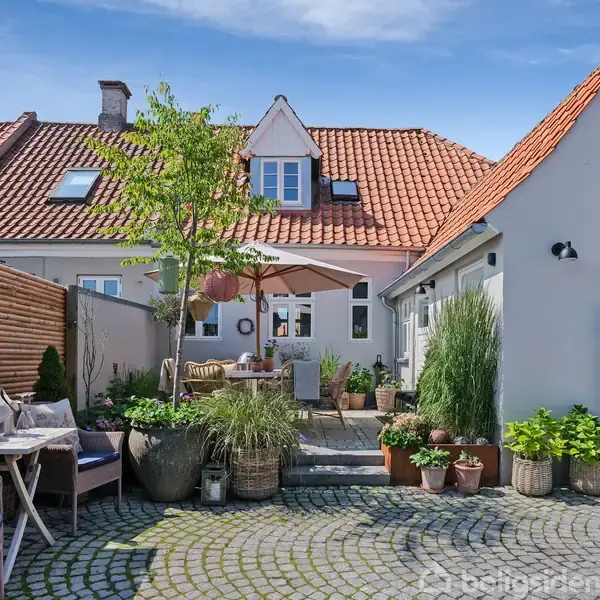 Rummeligt gårdhaveområde med møbler og flere planter på brostensbelægning. Ejendommen er omkranset af huse med røde tegltage. En parasol skaber skygge ved siddeområdet.