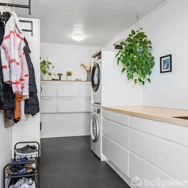 Et bryggers med hvide skabe og en vask, indeholder en vaskemaskine og tørretumbler stablet. Jakker hænger til venstre. Grønne planter og hylder med pynt skaber et hjemligt miljø.