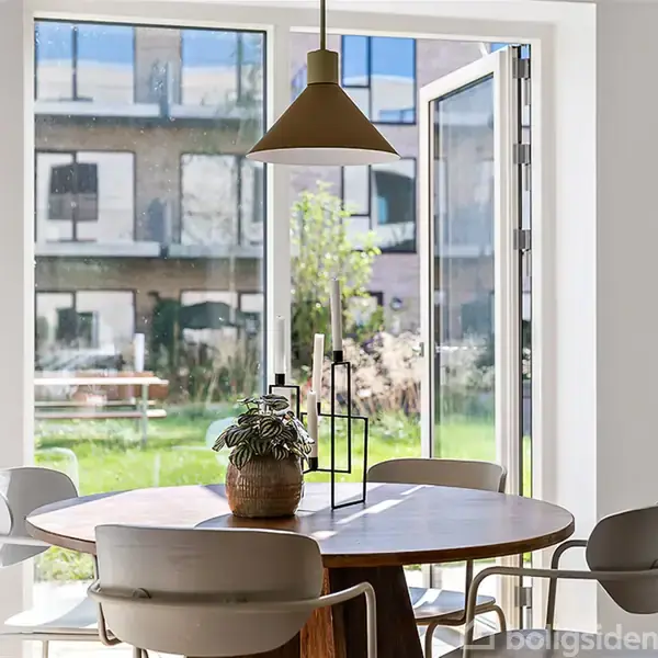 Et rundt spisebord med grå stole står i et lyst rum. Bordet har en grøn plante og lysestager. Store glasdøre åbner mod en have med græs og bygning i baggrunden.