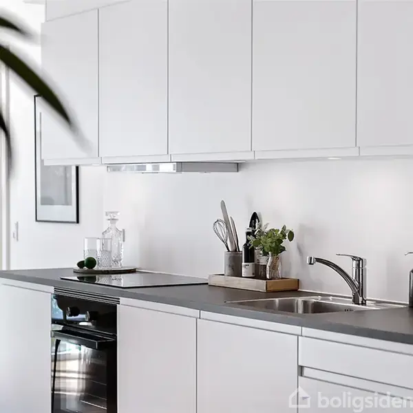 Køkkenskabe står over en disk, hvor glas og bestik hviler ved siden af en vask. Lyse hvide elementer dominerer køkkenet. En grøn plante er delvist synlig i forgrunden.