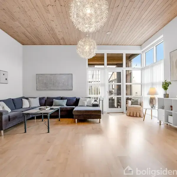 Grå sofa står i opholdsstue med stort vinduesparti. Lamper hænger fra træt loft; maleri og planter dekorerer rummet. Skandinavisk indretning med lyse farver og minimalistisk møblement.