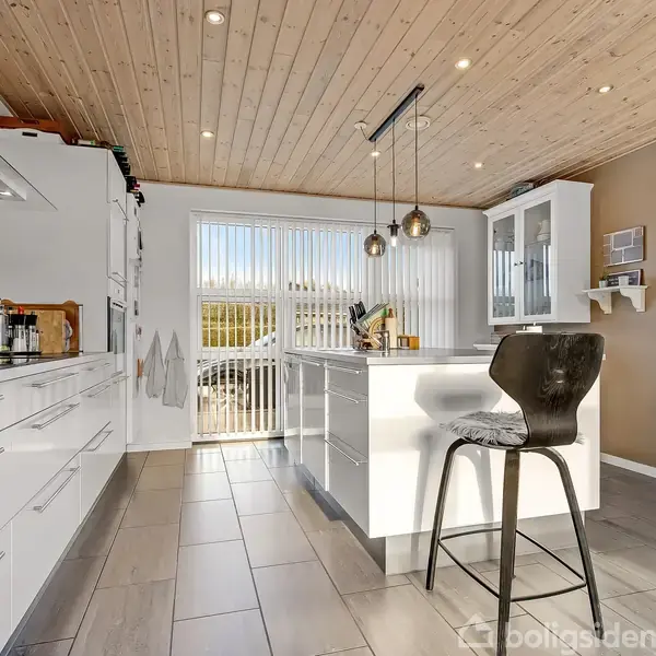 Køkkenø med sort barstol står i et lyst, moderne køkken. Loftet er af træ, og der er store vinduer bag øen. Hvide skabe og hylder omgiver rummet.