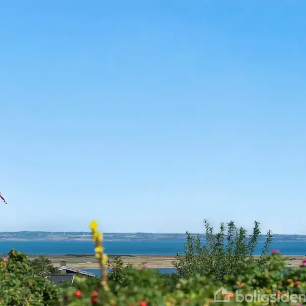 Flag drastisk vinkende i vinden, omkranset af grønne buske, med en blå havbaggrund og en lysblå himmel.