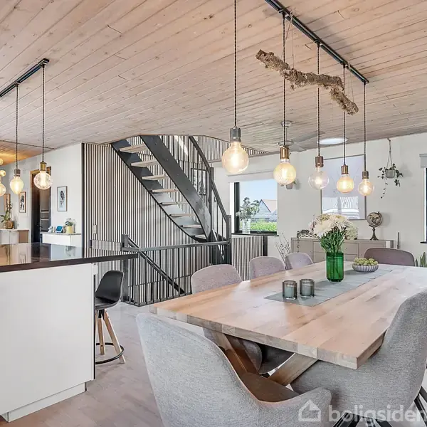 Køkken-alrum med spisebord og stole i forgrunden, under dekorative hængende lamper. Til venstre ses en køkkenø med barstole. En trappe leder til en overetage. Vinduer giver naturligt lysindfald.
