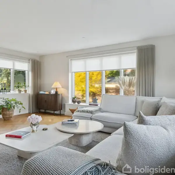 En stor, hvid sofa med puder står foran et sofabord i et lyst stueområde. Store vinduer med gardiner slipper sollys ind. Plakater og planter pynter rummet.