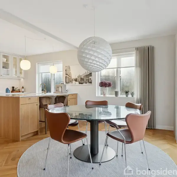 En rund glasbord er omgivet af fire brune stole i en lys spisestue. En stor, rund lampe hænger over bordet. Køkken med træelementer ses i baggrunden.