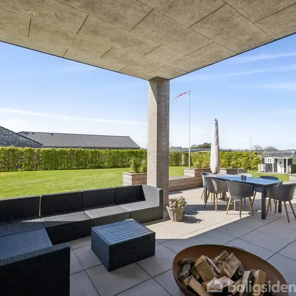 Terrasse med sort loungemøblering og bord med stole. I forgrunden en bålplads. Omgivet af en velplejet have med græsplæne og flagstang samt hæk. Klar blå himmel.