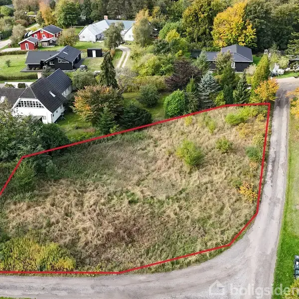 Ubebygget grund afgrænset med rød linje, dækket af vildtvoksende græs, omgivet af gader og huse i en frodig, skovklædt forstadsomgivelse.