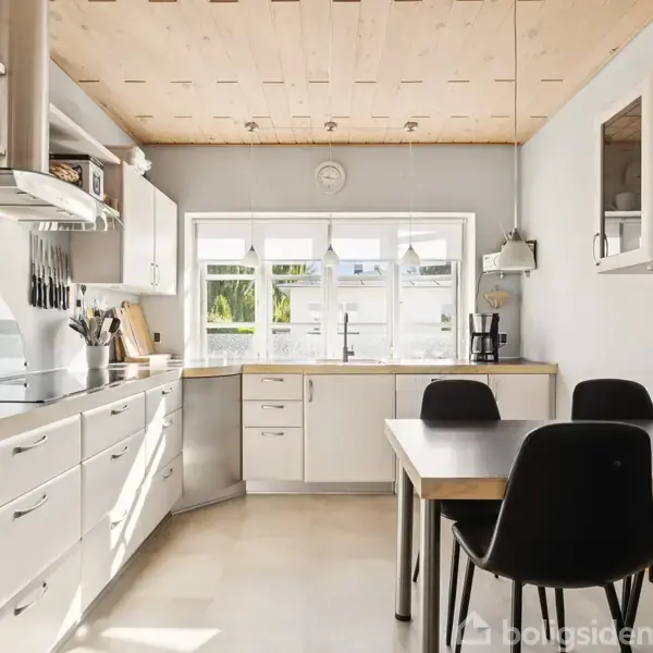 Et lyst køkken med hvide skabe og skuffer, indbyggede apparater og et spisebord med sorte stole. Store vinduer lader naturligt lys komme ind, med udsigt til have.