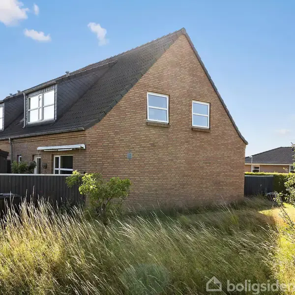 Et rødt murstenshus står stille, omgivet af højt græs og buske. Det har en sort tag og flere vinduer, med en lyseblå himmel og få skyer i baggrunden.