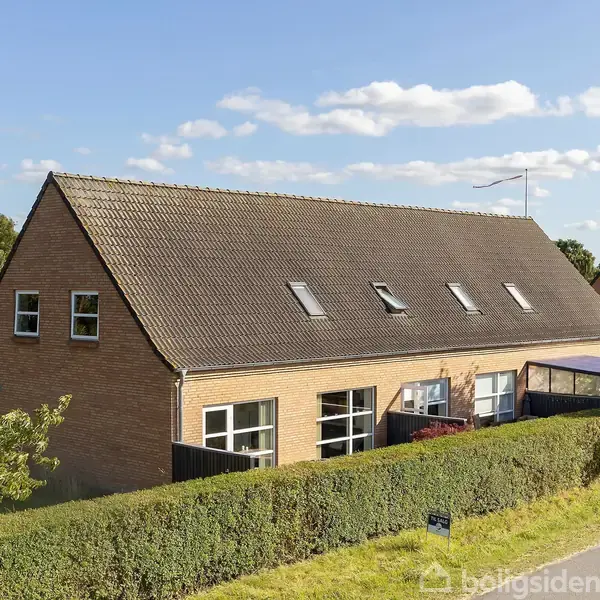 Et rødt murstenshus med skråt tag har flere tagvinduer. Det ligger i et grønt område med en hæk foran og en vej til højre.